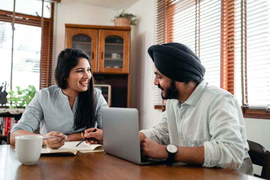 couple sitting at table smiling whilst talking, male is pointing to female's notepad whilst laptop is in front of male