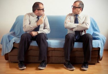 man speaking with angry facial expression towards another man sitting next to him on sofa