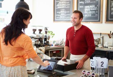 customer at counter of coffee shop using digital payment service device whilst smiling at shopkeeper